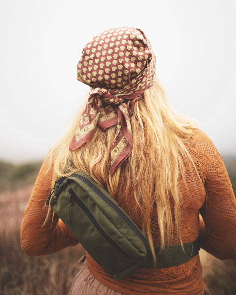 A woman wearing the Everyday Fanny Pack (Fern) in a lush outdoor setting, highlighted by her patterned scarf and hair.
