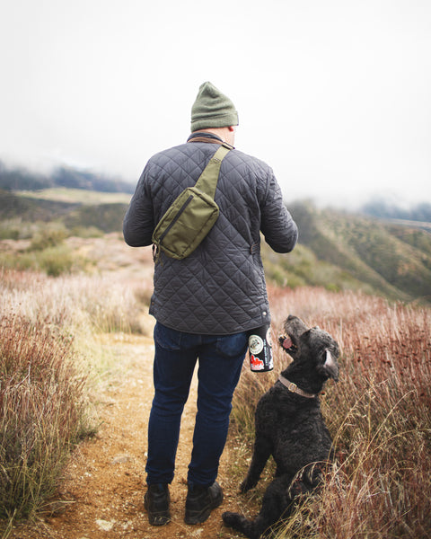 Person hiking with dog wearing Everyday Fanny Pack (Fern) as a crossbody sling, highlighting outdoor versatility and style.