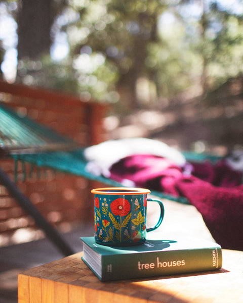 Garden Buzz Enamel Mug 16oz on a wooden table beside a book in a picturesque outdoor setting.
