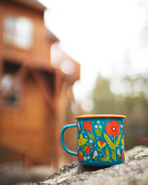 Garden Buzz Enamel Mug 16oz displayed outdoors with colorful floral design on a stone surface.