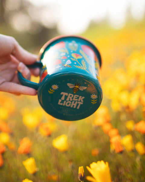 Colorful Trek Light Garden Buzz Enamel Mug 16oz in a field of orange flowers, perfect for outdoor adventures.
