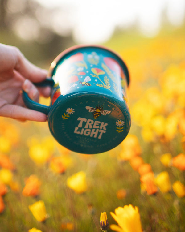 Colorful Trek Light Garden Buzz Enamel Mug 16oz in a field of orange flowers, perfect for outdoor adventures.