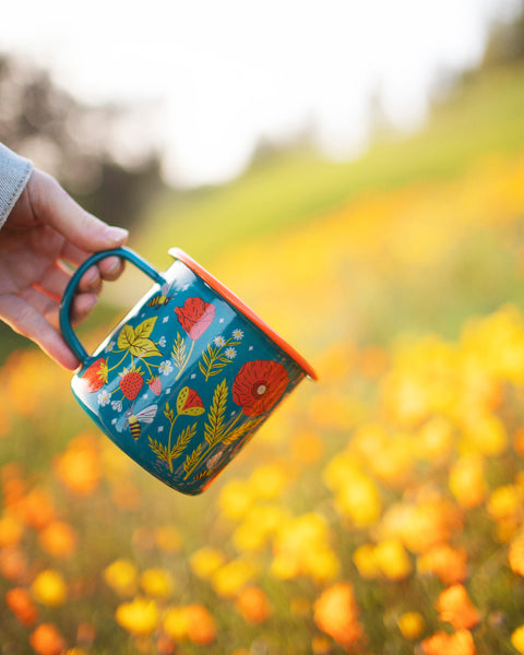 Garden Buzz Enamel Mug 16oz held in hand against a colorful flower background.