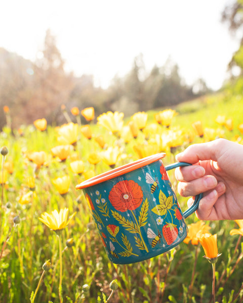 Garden Buzz Enamel Mug 16oz held in a field of yellow flowers, featuring vibrant floral design. Perfect for your next Trek Light adventure.