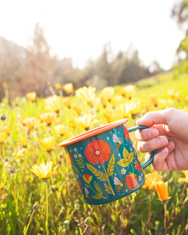 Garden Buzz Enamel Mug 16oz held in a field of yellow flowers, featuring vibrant floral design. Perfect for your next Trek Light adventure.