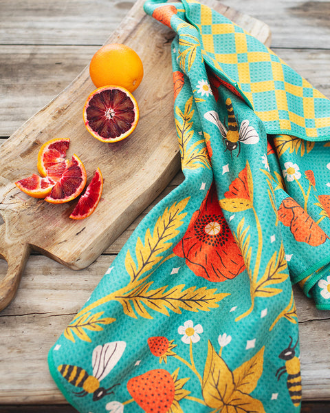 Garden Buzz Kitchen & Tea Towel on a wooden board with oranges and vibrant designs of fruits and bees.