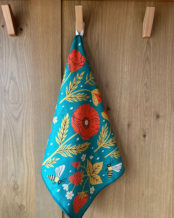 Eco-friendly Garden Buzz kitchen dish towel with vibrant floral and bee design, hanging on a wooden hook. Super absorbent.