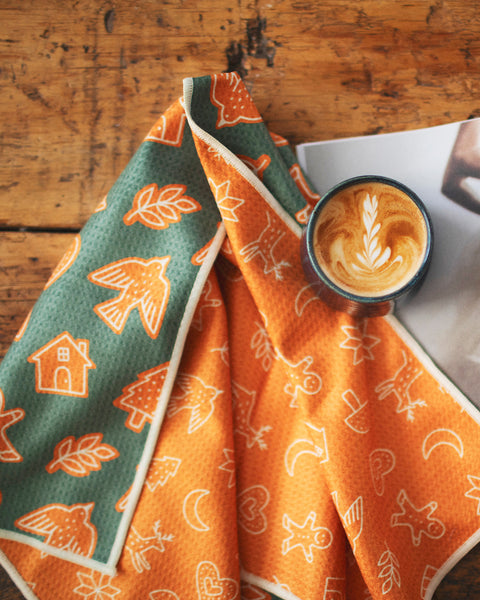 Gingerbread Kitchen & Tea Towel folded next to a coffee mug on a wooden table, showcasing festive designs.