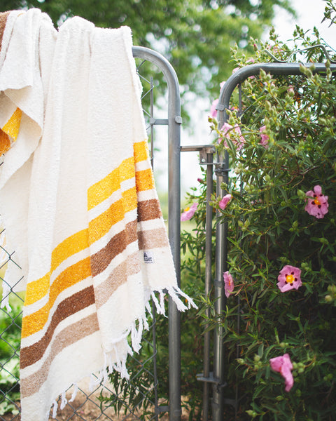 Golden Age Blanket hanging on a fence surrounded by flowers, showcasing its lightweight and colorful striped design.