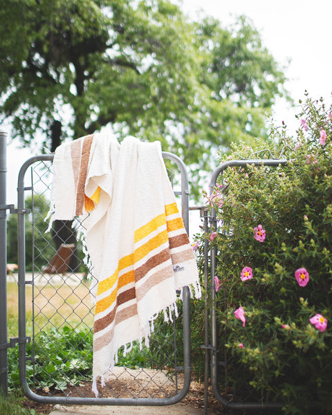 Golden Age Blanket hanging on a metal fence surrounded by greenery and flowers, showcasing its striped design.