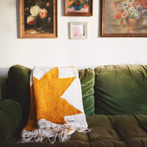 Cozy Goldie blanket handwoven from recycled clothing materials, draped over a green couch, showcasing a beautiful geometric design. By Trek Light.