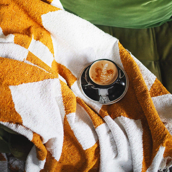 Cozy handwoven Goldie blanket in geometric design, made from recycled clothing materials, with a cup of coffee on top. By Trek Light.