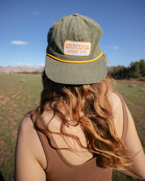 Woman wearing Grateful Every Day Trek Light corduroy snapback hat, enjoying nature with a green field background.