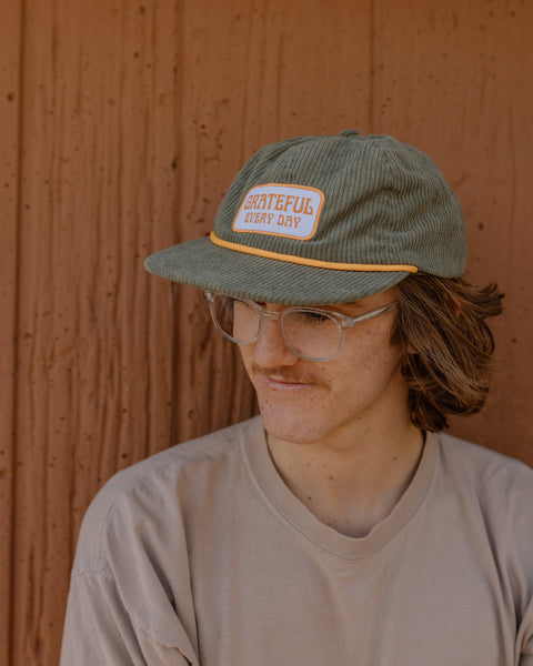 Man wearing Grateful Every Day hat by Trek Light, green corduroy snapback with flat brim, showcasing gratitude style.
