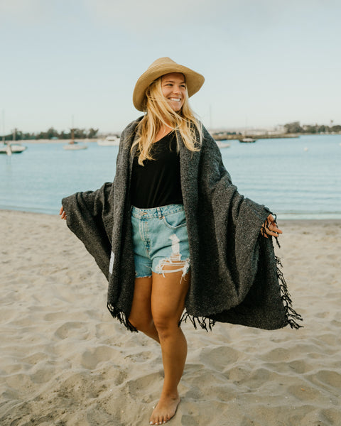 Woman wearing a Greyson Blanket at the beach, showcasing its stylish, handwoven design.