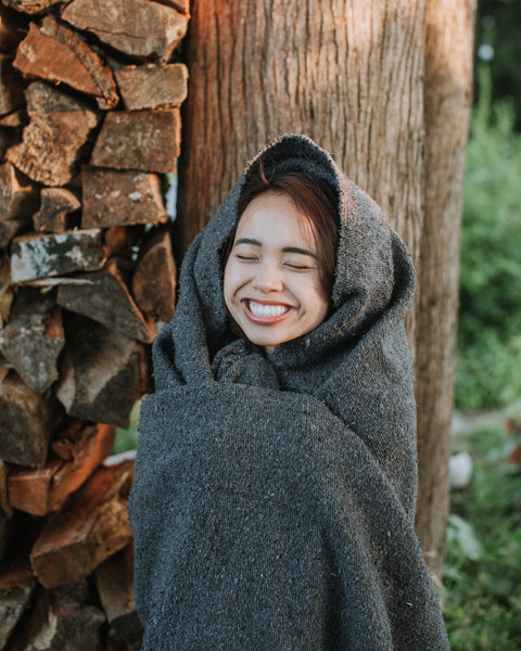 Happy woman wrapped in a Greyson Blanket near stacked firewood in a lush garden.