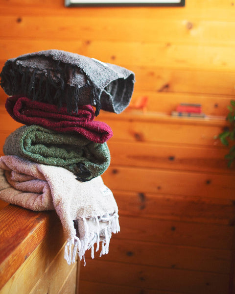 Stack of cozy blankets including the Greyson Blanket in a rustic wood setting.