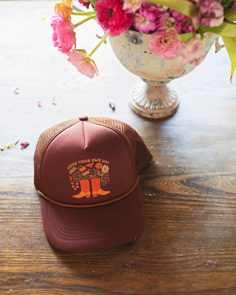 Grow Your Own Way Hat displayed on a wooden table with flowers, featuring a vibrant design and adjustable fit.