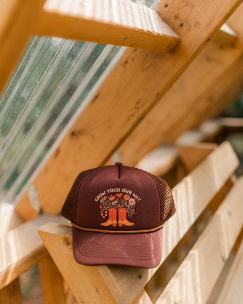Grow Your Own Way Hat displayed on wooden shelf in greenhouse, featuring colorful design with flowers and boots.