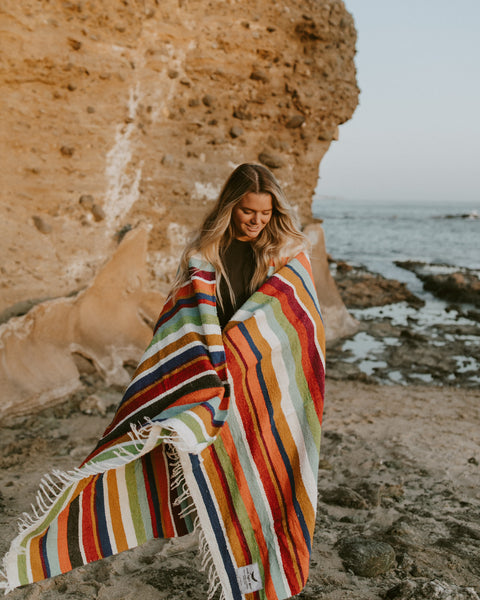 Woman wrapped in striped Halley's Comet Blanket at the beach, enjoying the view.