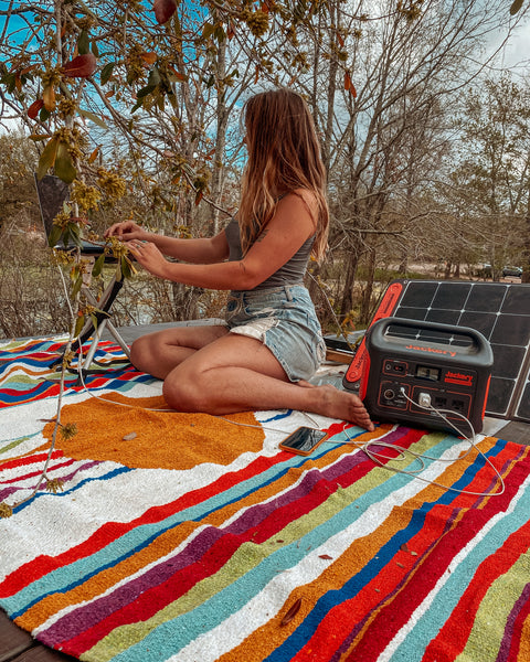 Woman working on a laptop outdoors, using a solar power generator, sitting on a colorful striped blanket.