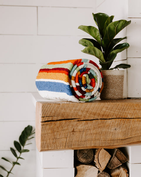 Trek Light's Halley's Comet Blanket, vibrantly striped and handwoven, rolled up beside a plant on a wooden shelf.