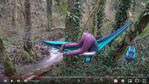 Person humorously falls out of hammock in forest, overlooking a creek, with backpack hanging nearby.