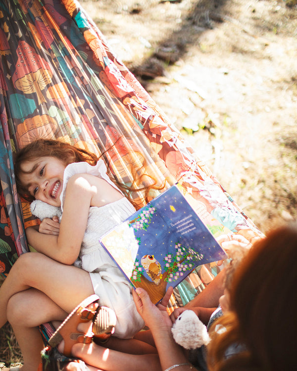 A joyful child reading a book while relaxing with a parent in a Double Hammock & Pro Hanging Straps (Midnight Mushroom Print).