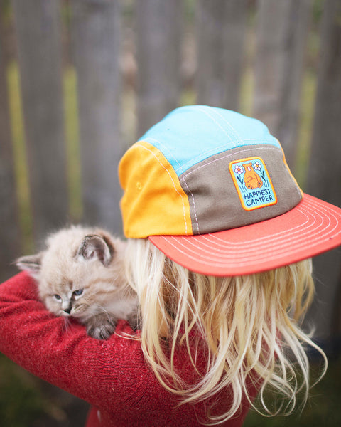 Child wearing a Happiest Camper Kids Hat while holding a kitten outdoors.
