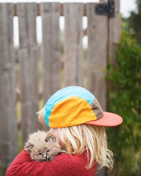 Child wearing a colorful Happiest Camper Kids Hat while holding a kitten, ideal for outdoor adventures.