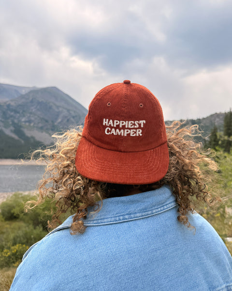 Happiest Camper Hat worn outdoors with mountains in the background, showcasing the embroidered design.