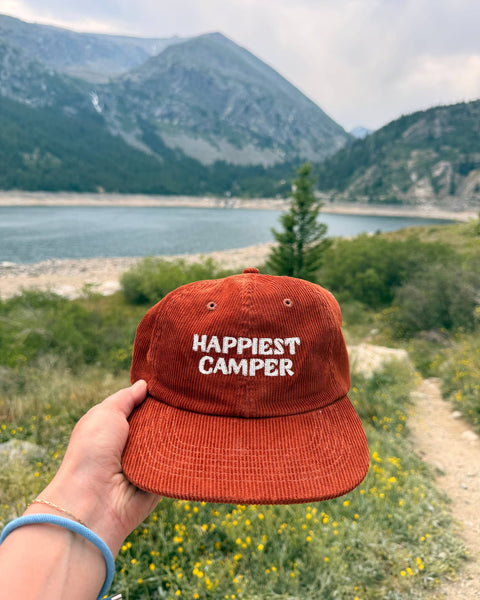 Happiest Camper Hat displayed with a scenic mountain backdrop and greenery, perfect for camping and outdoor adventures.