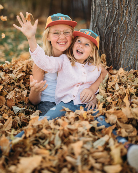 Two kids wearing Happiest Camper Kids Hat, playing in leaves and smiling, perfect for outdoor fun and gifting.