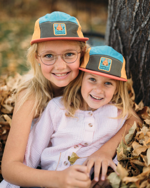 Two smiling girls wearing the Happiest Camper Kids Hat, playing in autumn leaves.