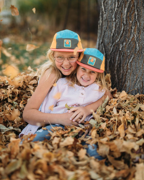 Two smiling children wearing Trek Light hats, sitting in a pile of autumn leaves near a tree. Perfect moment of outdoor joy.