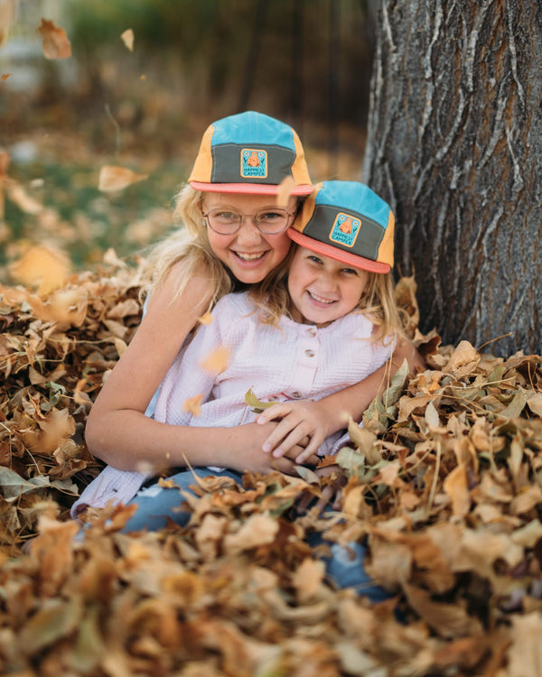 Two smiling children wearing Trek Light hats, sitting in a pile of autumn leaves near a tree. Perfect moment of outdoor joy.