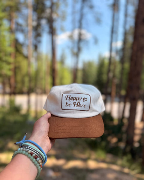 Happy To Be Here Hat held in hand, featuring a brown brim and comfortable design, perfect for all-day wear.