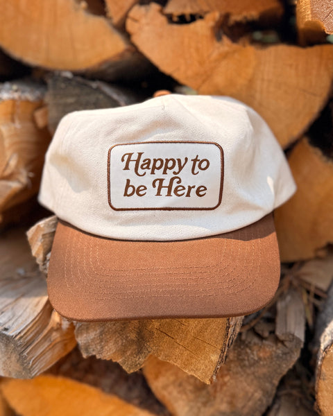 Happy To Be Here Hat resting on wooden logs with a captivating design, ideal for style and comfort.