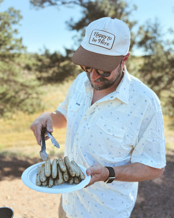 A man wearing a Happy To Be Here Hat holds a plate of sausages outdoors.