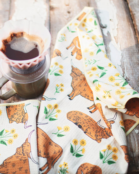 Highland Cows Kitchen & Tea Towel displayed with a mug of coffee on a wooden table.
