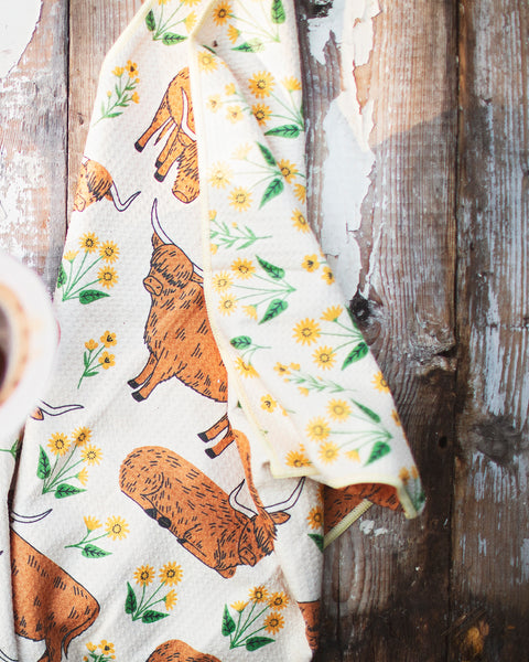 Highland Cows Kitchen & Tea Towel with vibrant designs and floral patterns on a rustic wooden table.