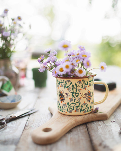 Honey Bee Enamel Mug 16oz filled with flowers on a wooden surface, perfect for adding charm to your mornings.