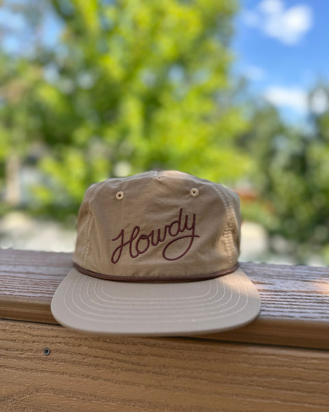 Howdy Hat displayed on a wooden surface with greenery in the background, featuring a flat-brim and adjustable fit.