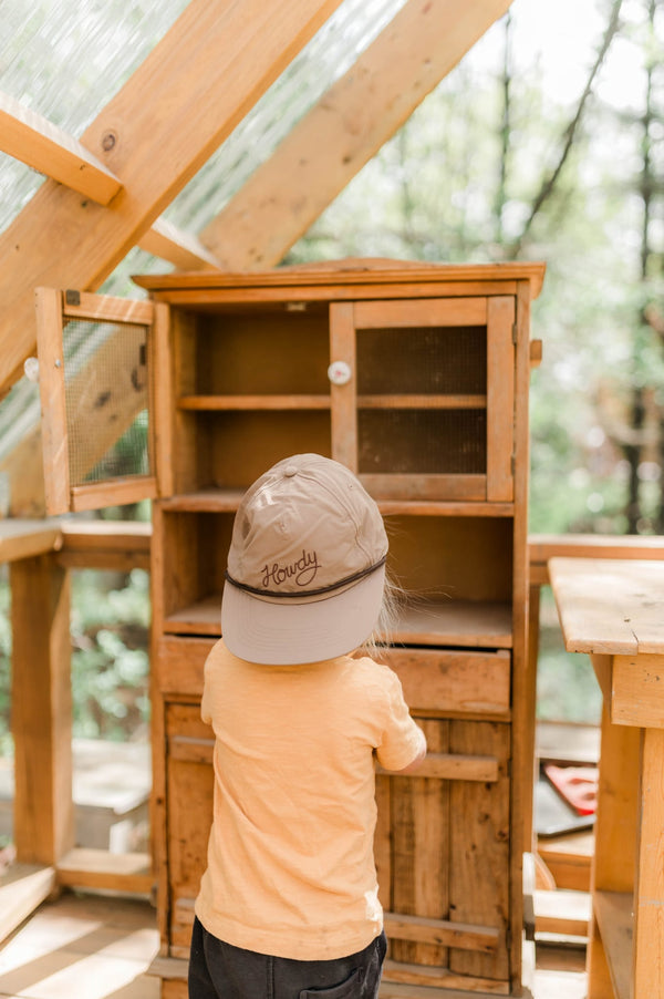 Child exploring wooden cabinet wearing Howdy Kids Hat, showcasing fun and adventure in outdoor play.