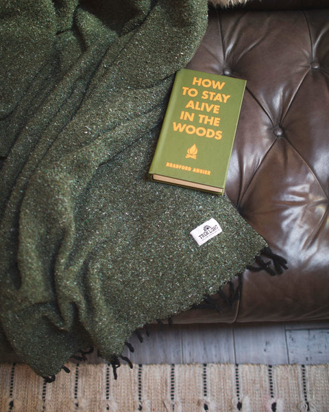 Cozy Humboldt Blanket draped over a leather couch with 'How to Stay Alive in the Woods' book, handwoven and eco-friendly design.