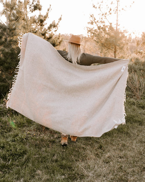 Woman holding a sand-colored Huntington Blanket in nature, perfect for beach days and cozy nights. Trek Light adventure vibes.