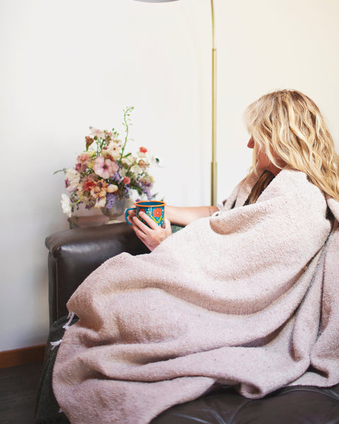 A woman wrapped in a Huntington Blanket, holding a colorful mug while relaxing on a couch with flowers nearby.