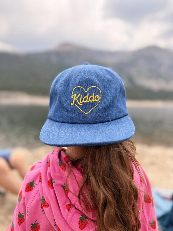 A person viewed from behind wearing a blue denim-style baseball cap with yellow embroidered text reading 'Kiddo' inside a heart outline, along with a pink strawberry-patterned top, set against a blurred outdoor mountain background.