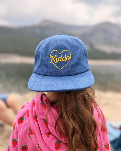 A person viewed from behind wearing a blue denim-style baseball cap with yellow embroidered text reading 'Kiddo' inside a heart outline, along with a pink strawberry-patterned top, set against a blurred outdoor mountain background.