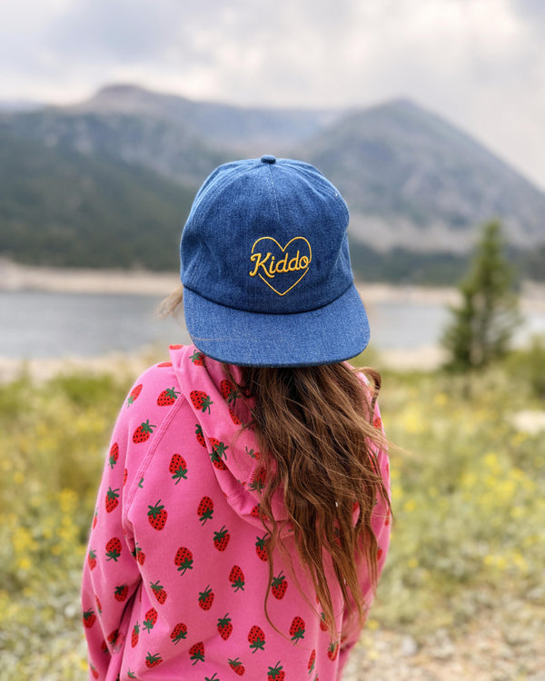 A child viewed from behind wearing a blue denim baseball cap with 'Kiddo' embroidered in yellow inside a heart, paired with a pink strawberry-patterned hoodie, set against a blurred mountain and lake background.